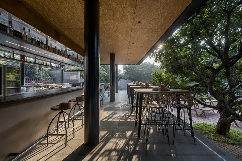 Covered terrace with cantilevered textured soffit, bar counter, and high tables casting shadows beside planted trees