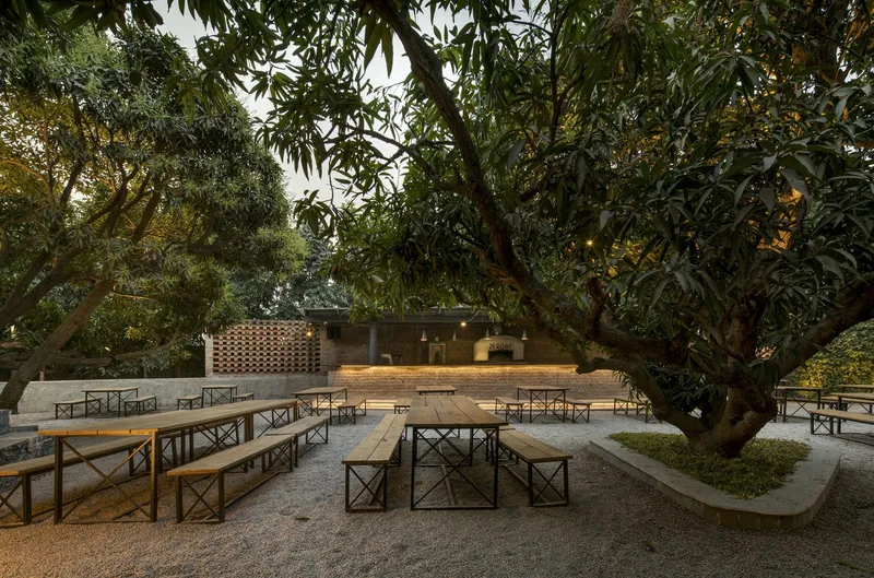 Outdoor dining area with timber benches beneath mature trees and perforated brick wall beyond