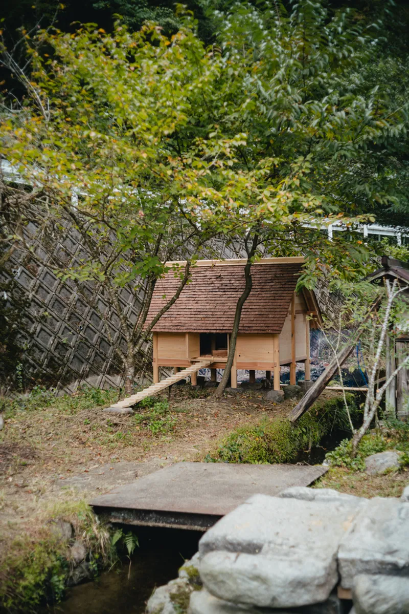 Elevated timber structure with a cedar shingle roof beside a stone-edged water channel in autumn