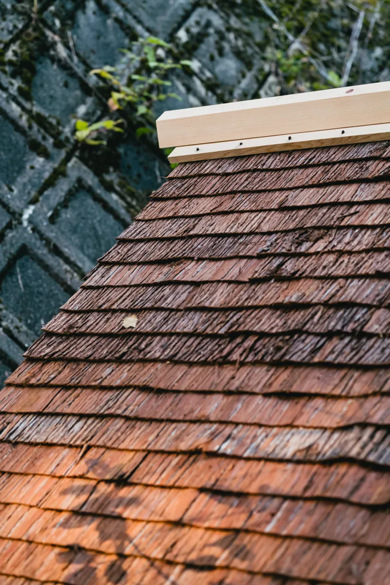 Cedar shingle roof with light timber fascia board against a textured stone wall backdrop