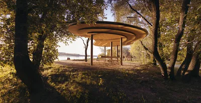 This architectural image showcases a modern pavilion nestled within a park-like setting. The structu