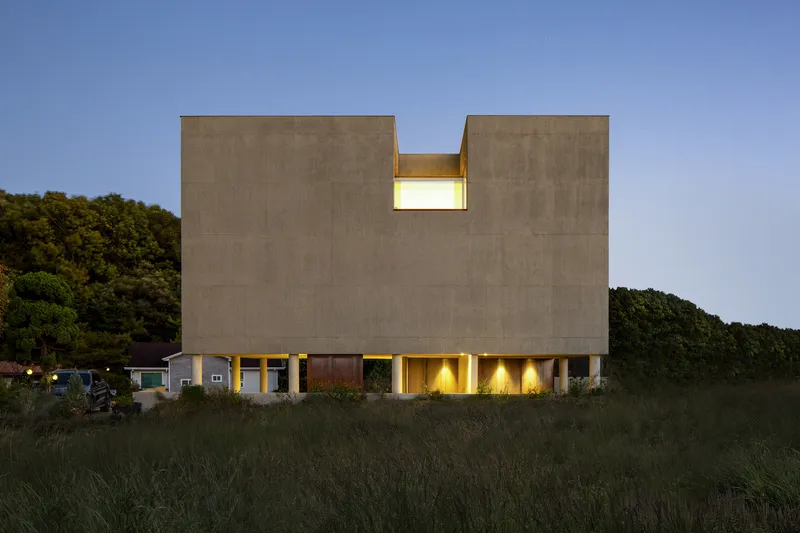 Notched concrete block facade with illuminated base and recessed window at dusk in coastal grassland