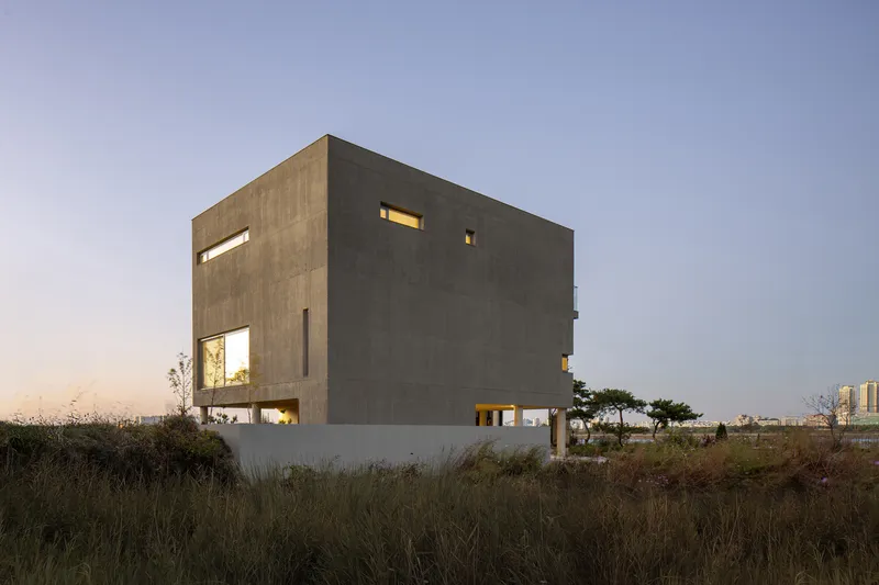 Cantilevered concrete cube with scattered window openings above a lit colonnade at twilight
