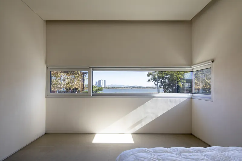 Bedroom with horizontal ribbon window framing distant water and sunlight casting shadows on polished floor