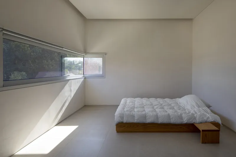 Bedroom with angled clerestory window and horizontal ribbon window framing sunlight across concrete floor