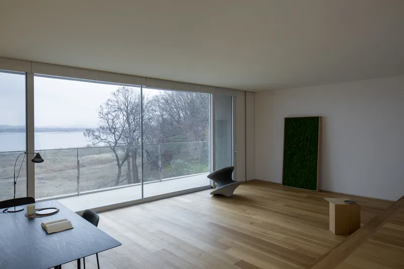 Living room with full-height glazing overlooking bare trees and water under overcast sky