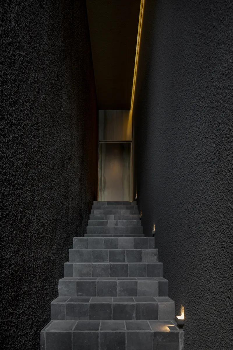 Stone staircase ascending between black textured walls with a narrow vertical slot of warm light above