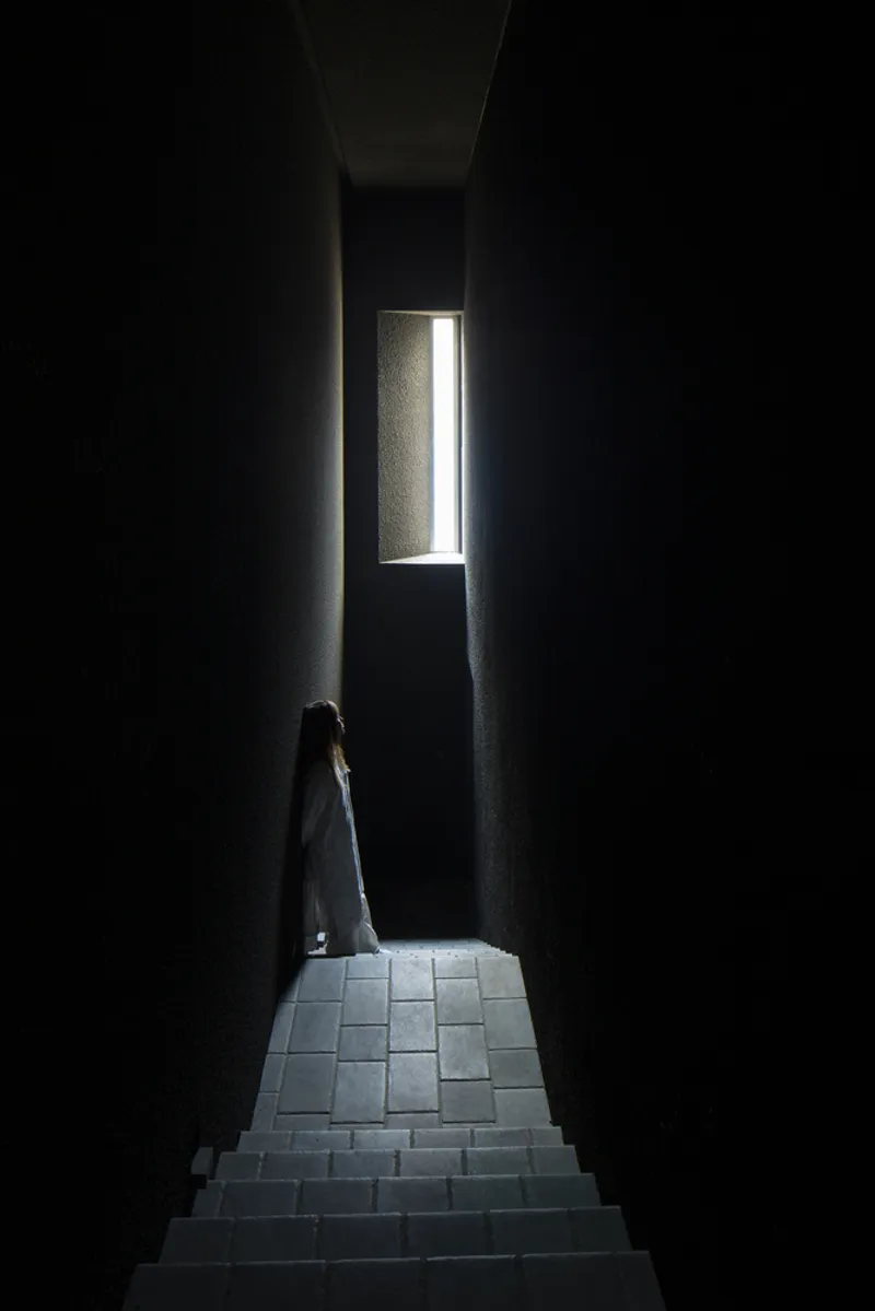 Figure standing on stone stairs in a dark passage illuminated by a single vertical window at the landing