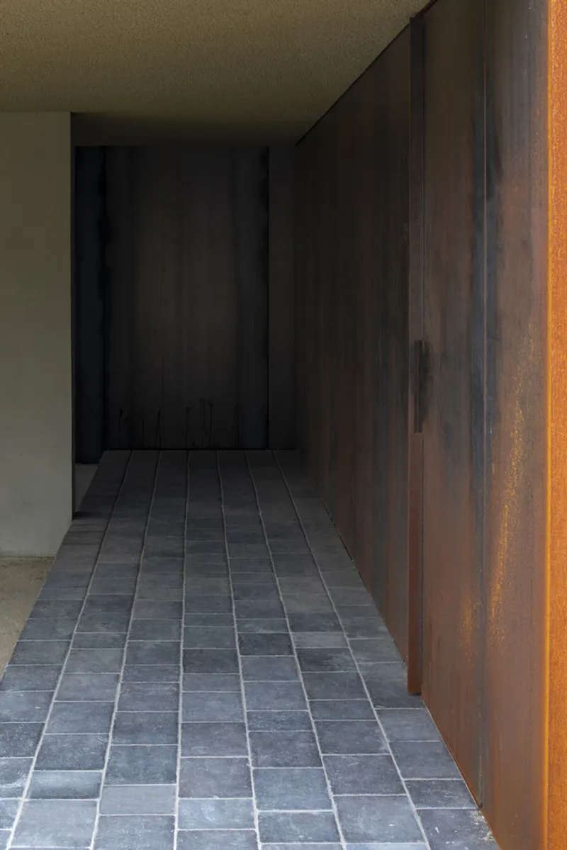 Narrow corridor with grey stone tile floor running between weathered steel wall panels toward a shadowed recess