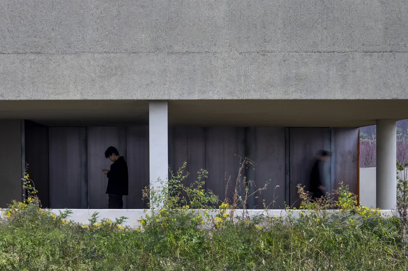 Exterior loggia with dark steel panels and two figures passing behind a low wildflower planting bed