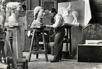 This black and white photograph captures a sculptor in his studio, working on a bust. The studio is 