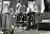 This black and white photograph captures a sculptor in his studio, working on a bust. The studio is 