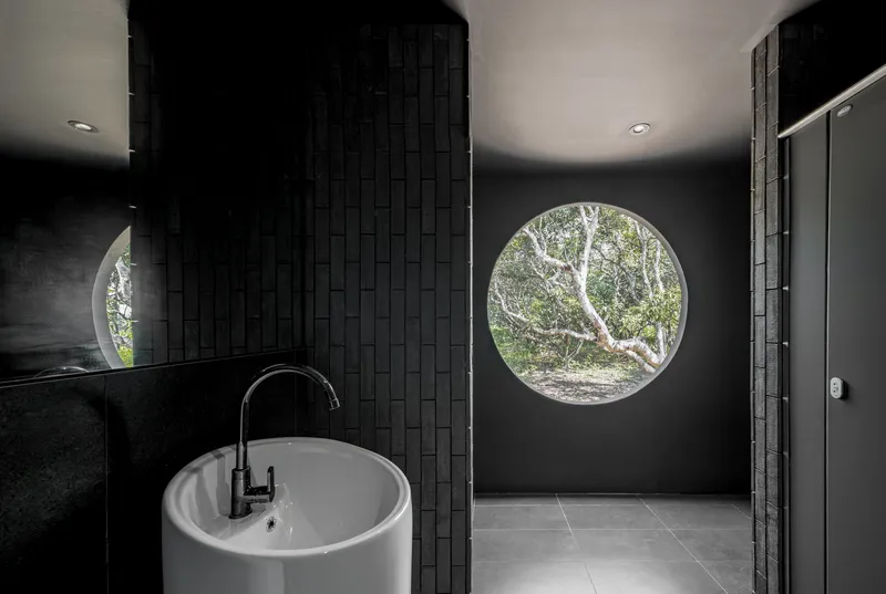 Dark-tiled bathroom with a round picture window framing the mango trees