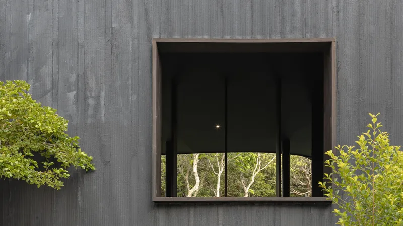 Deep-set square window punched into a charred-timber facade with leaves in the foreground
